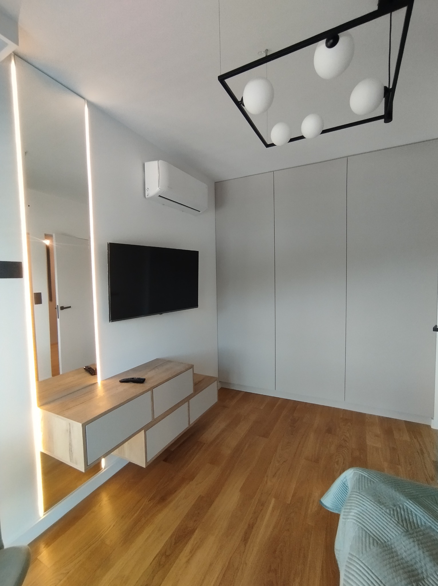 Nowoczesna sypialnia z dużą szafą w zabudowie, podświetlanym lustrem, wiszącą szafką RTV z szufladami i drewnianą podłogą, oświetlona lampą sufitową z białymi kulami.
