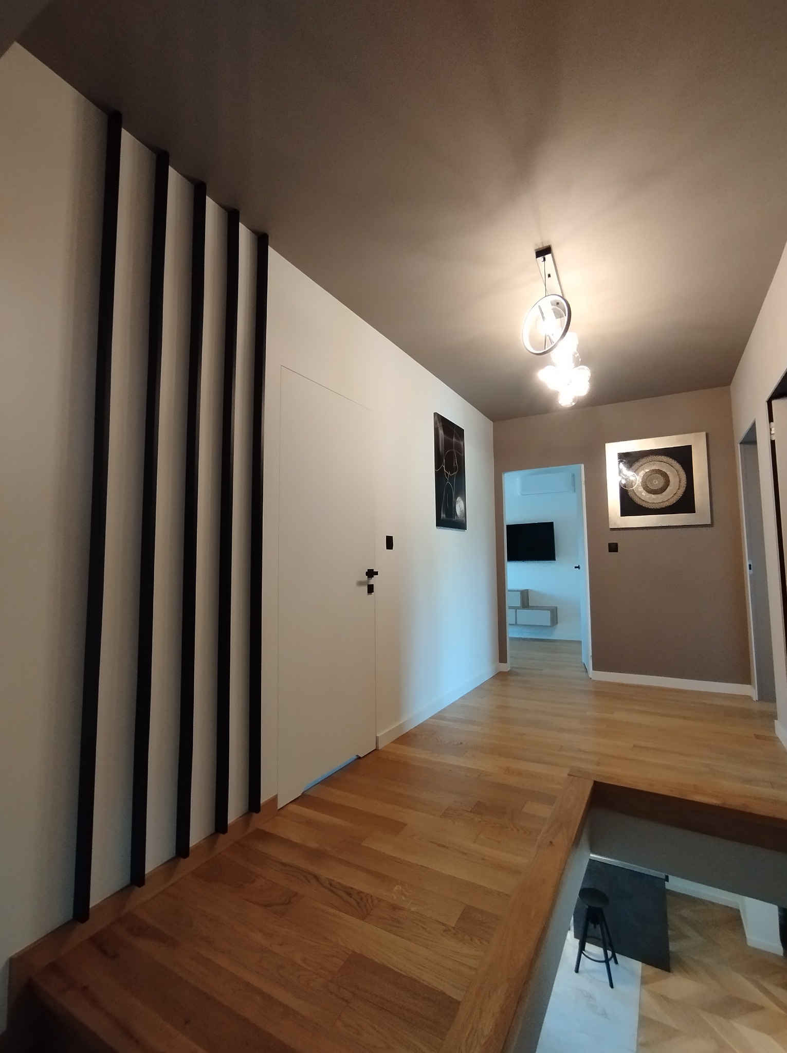 Nowoczesny przedpokój z drewnianą podłogą, czarnymi listwami dekoracyjnymi na ścianie, białymi drzwiami, oświetleniem sufitowym i obrazami na ścianach w odcieniach beżu i bieli.