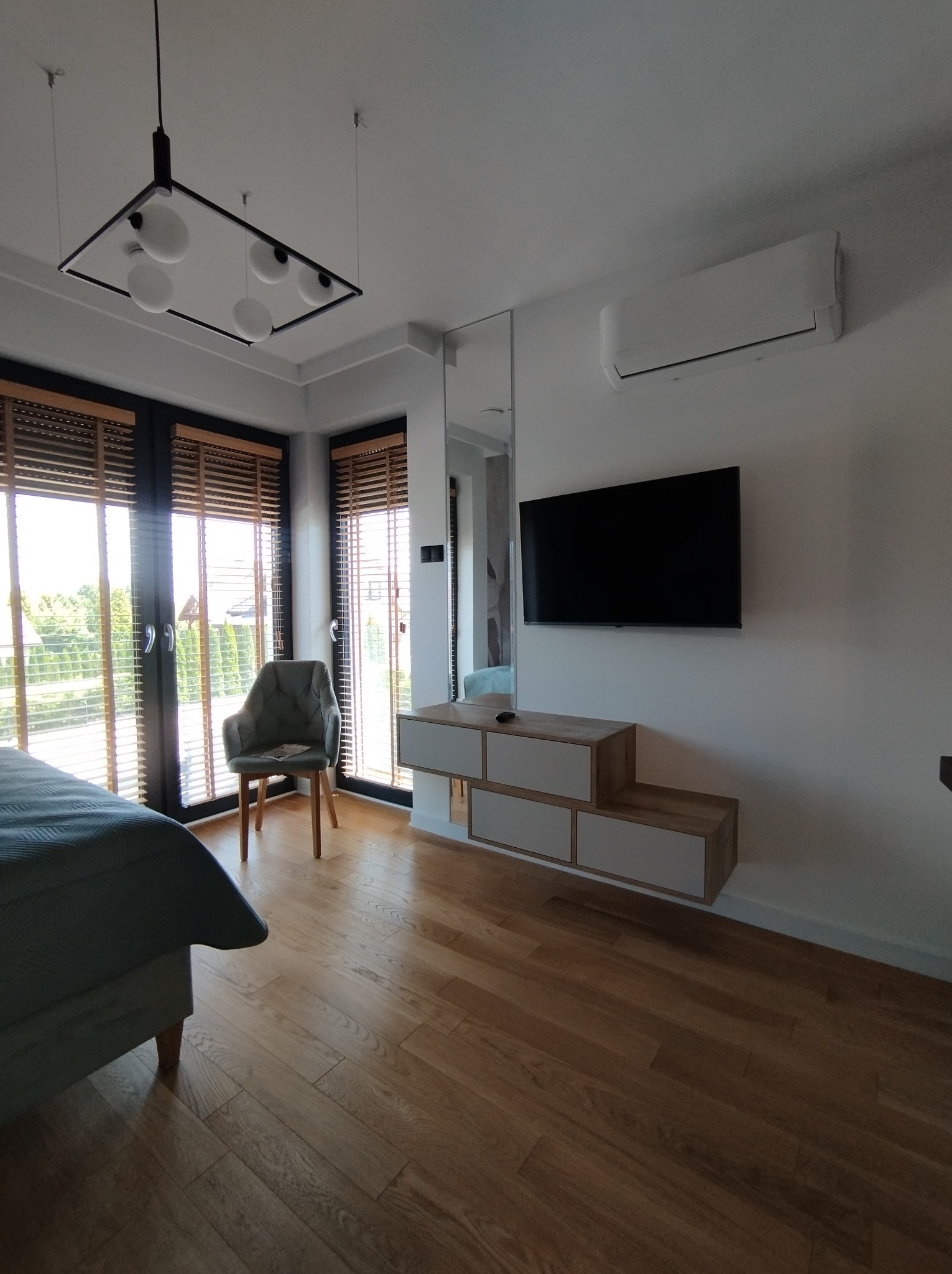 Nowoczesna sypialnia z drewnianą podłogą, łóżkiem z szarą pościelą, krzesłem obok okna z żaluzjami, wiszącą szafką RTV pod telewizorem i designerską lampą sufitową.