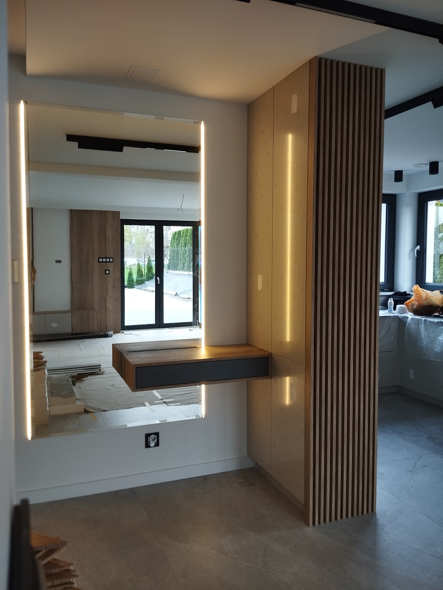 Nowoczesna szafka z lustrem i oświetleniem LED, w tle widoczna zabudowa kuchenna i okno tarasowe, podłoga w trakcie remontu.
