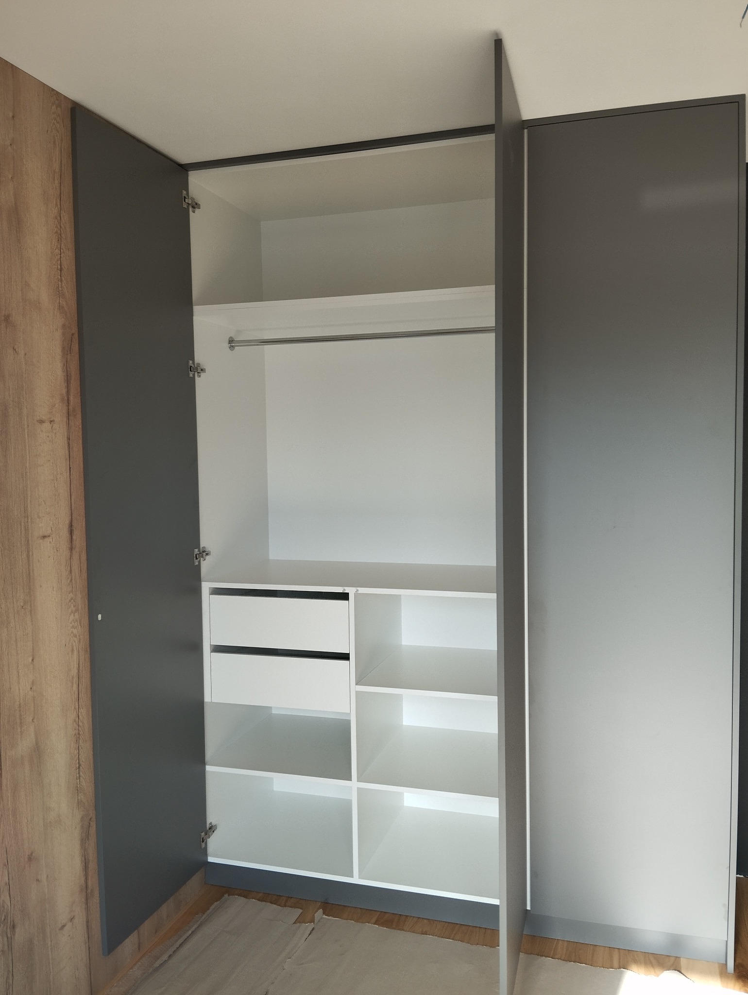 Otwarta szafa wnękowa z szarymi drzwiami i białym wnętrzem, widoczne półki, szuflady i drążek na ubrania, zarys podłogi zabezpieczony kartonem.