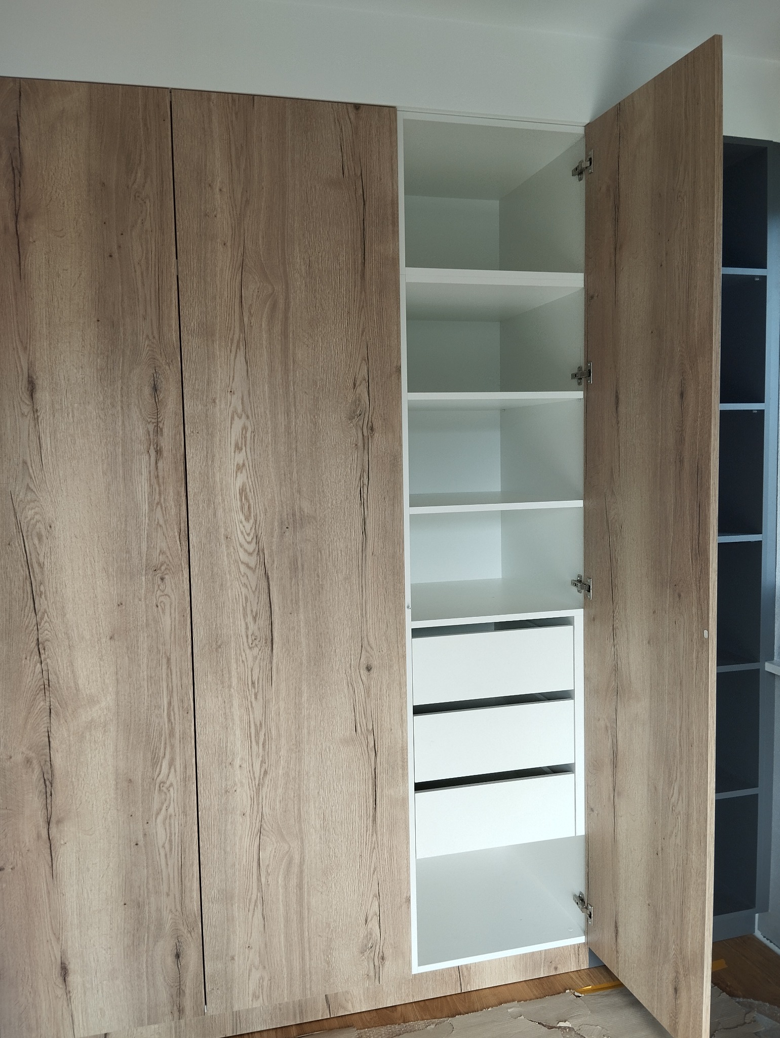 Wysoka szafa z drewnopodobnymi frontami, otwarte drzwi ukazują białe półki i trzy szuflady. Widoczne zawiasy i fragment podłogi z resztkami po montażu.