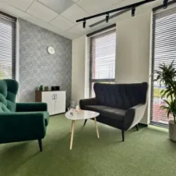 Jasne wnętrze gabinetu z zielonym fotelem typu uszak, szarą sofą, białym stolikiem kawowym i rośliną doniczkową przy oknie z żaluzjami.