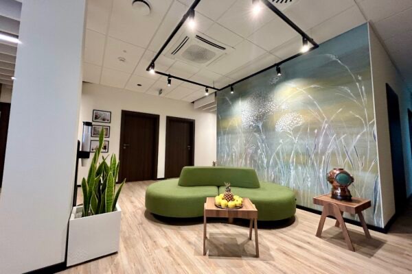 Wnętrze poczekalni: okrągła, zielona sofa, stolik z owocami, dekoracyjna ściana z motywem roślinnym, oświetlenie punktowe i donica z rośliną.