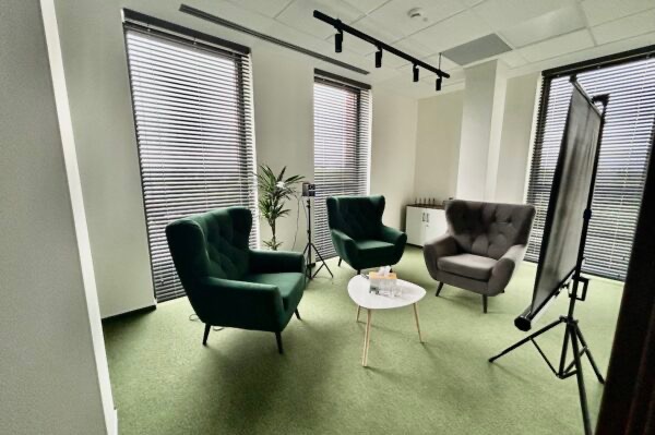 Jasne wnętrze gabinetu z trzema fotelami w odcieniach zieleni i szarości, stolikiem kawowym z wodą, roletami w oknach i lampą sufitową.