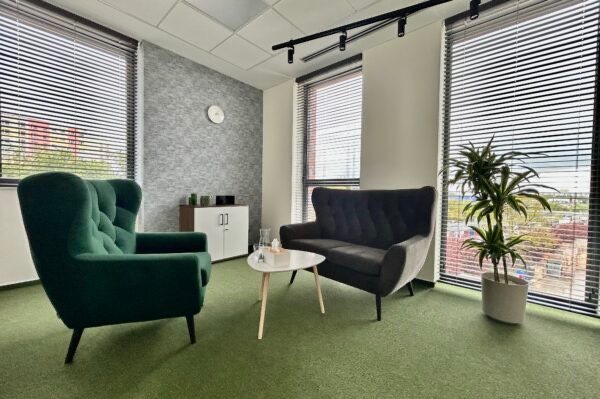 Jasne wnętrze gabinetu z zielonym fotelem typu uszak, szarą sofą, białym stolikiem kawowym i rośliną doniczkową przy oknie z żaluzjami.