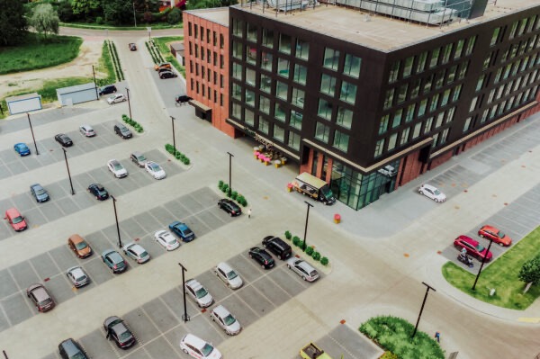 Widok z góry na parking przed nowoczesnym budynkiem biurowym z ceglaną elewacją i dużymi oknami; zaparkowane samochody różnych kolorów i modeli.