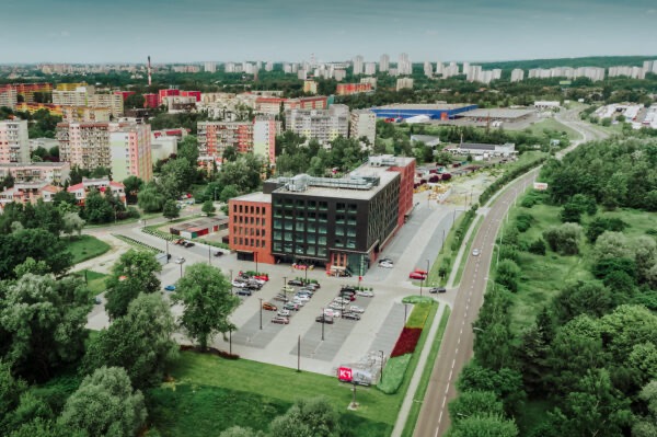 Widok z lotu ptaka na nowoczesny budynek biurowy z parkingiem i zielenią, otoczony zabudową miejską i drzewami.