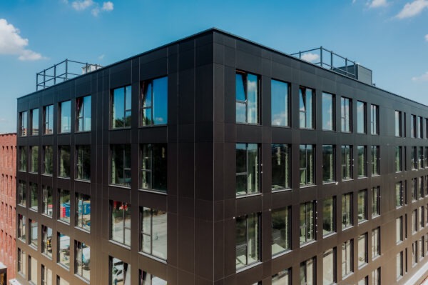 Nowoczesny, czteropiętrowy budynek biurowy z czarną elewacją z paneli kompozytowych i regularnym układem okien odbijających błękitne niebo.
