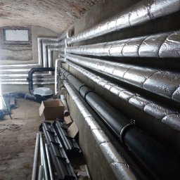 GrafTherm Usługi Sanitarne Leszek Graf - Instalacja rur w piwnicy z ceglanym sklepieniem. Rury izolowane srebrną folią, niektóre czarne, prowadzone wzdłuż ściany. Na podłodze narzędzia i materiały budowlane.