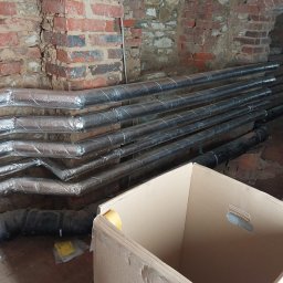 GrafTherm Usługi Sanitarne Leszek Graf - Rury izolowane folią aluminiową ułożone w piwnicy z ceglanym sklepieniem, obok kartonowe pudło. Widoczne złącza rur. Prace instalacyjne w trakcie realizacji.