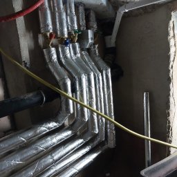 GrafTherm Usługi Sanitarne Leszek Graf - Izolowane rury instalacji wodnej z zaworami w piwnicy, widoczne elementy mocowania do sufitu. Żółty kabel wzdłuż rur. Instalacja po remoncie.