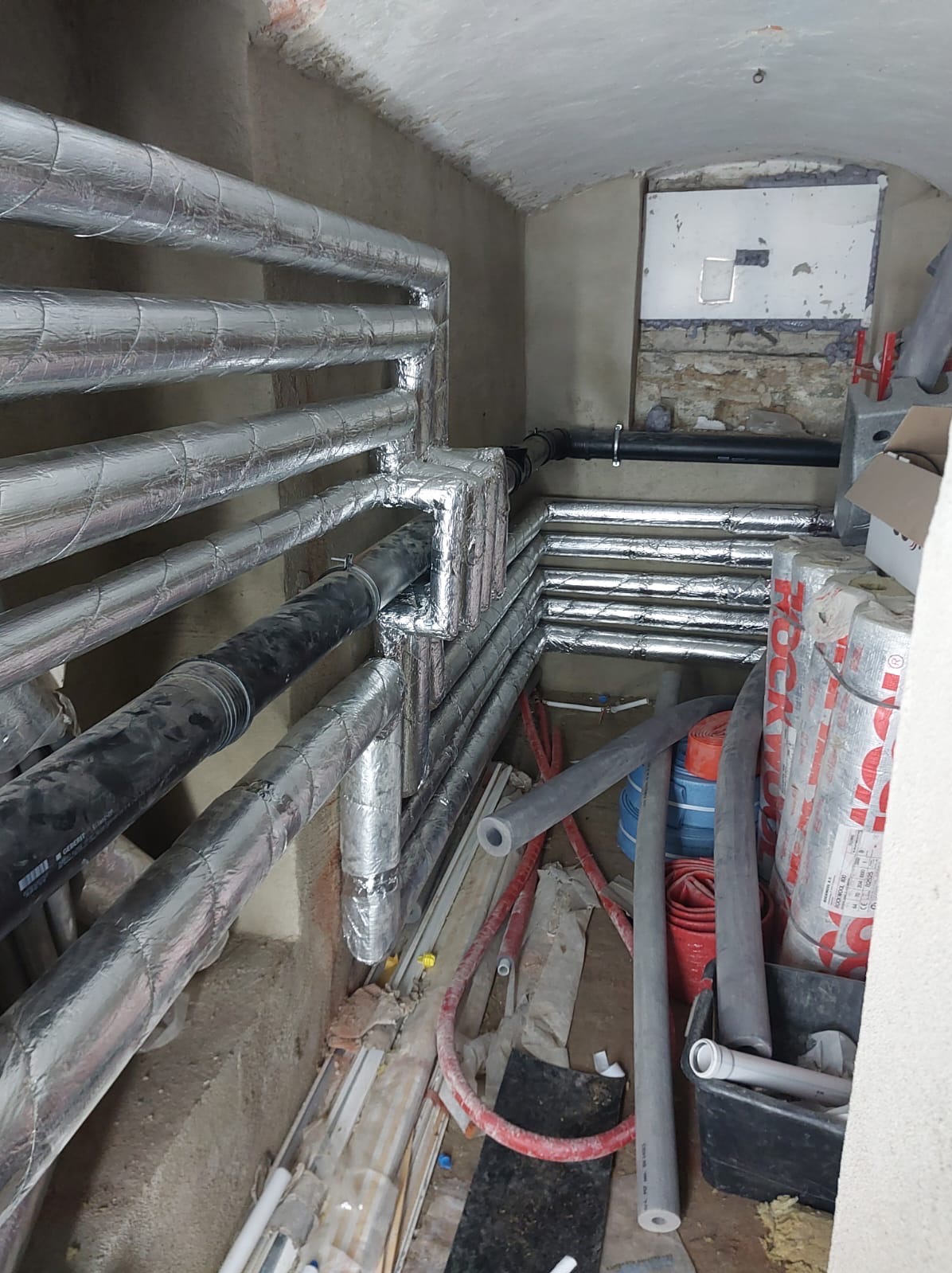 Izolowane rury w piwnicy, widoczne materiały izolacyjne Rockwool, instalacja grzewcza w trakcie montażu, rury czarne i srebrne, surowy stan wykończenia.