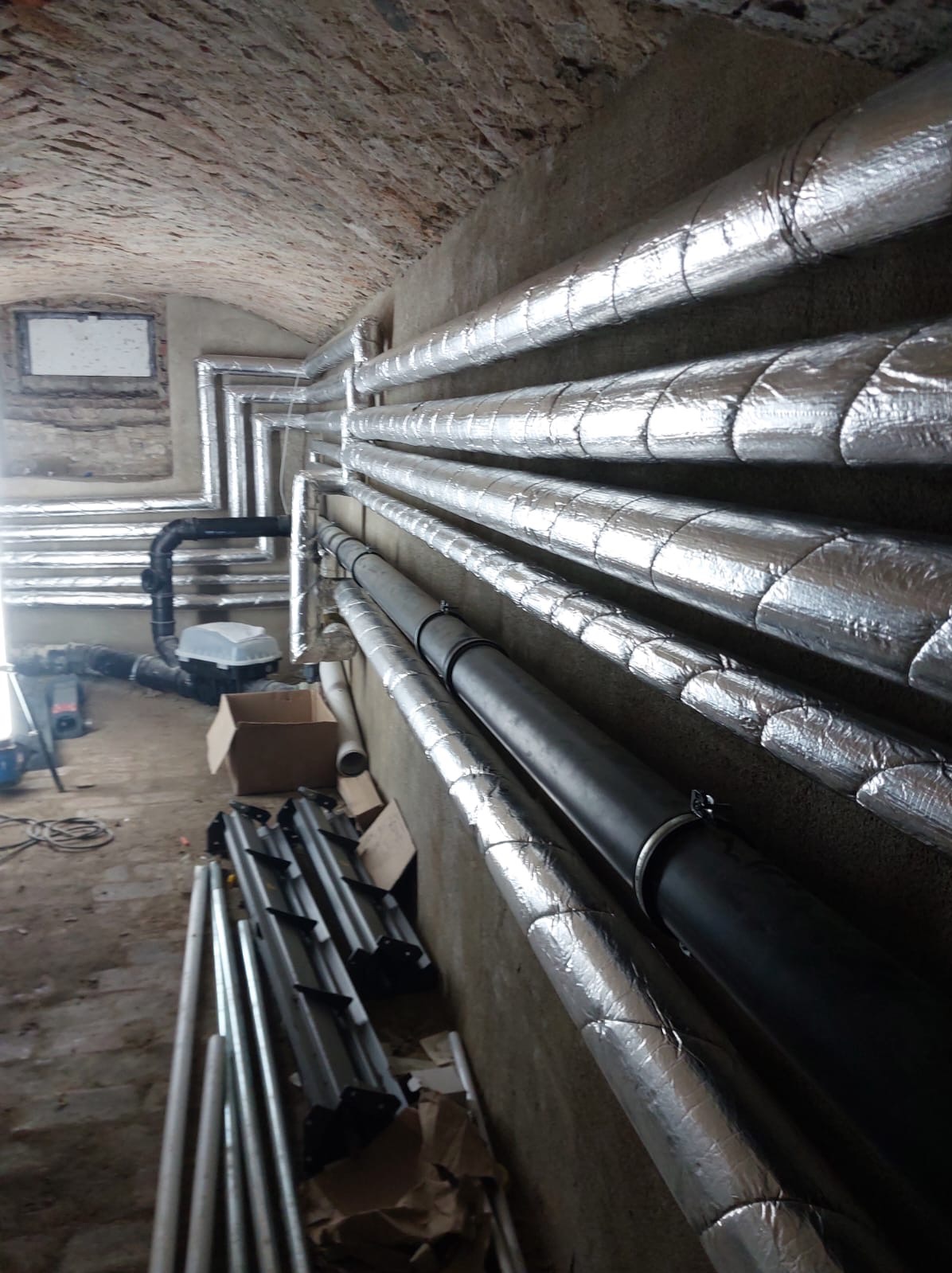 Instalacja rur w piwnicy z ceglanym sklepieniem. Rury izolowane srebrną folią, niektóre czarne, prowadzone wzdłuż ściany. Na podłodze narzędzia i materiały budowlane.