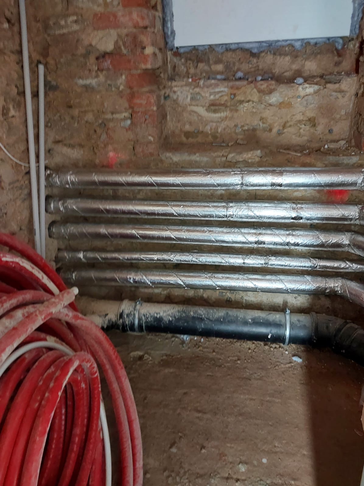 Ocieplone rury instalacji grzewczej ułożone w wykopie przy ceglanej ścianie. Czerwone rury ogrzewania podłogowego w rogu kadru.