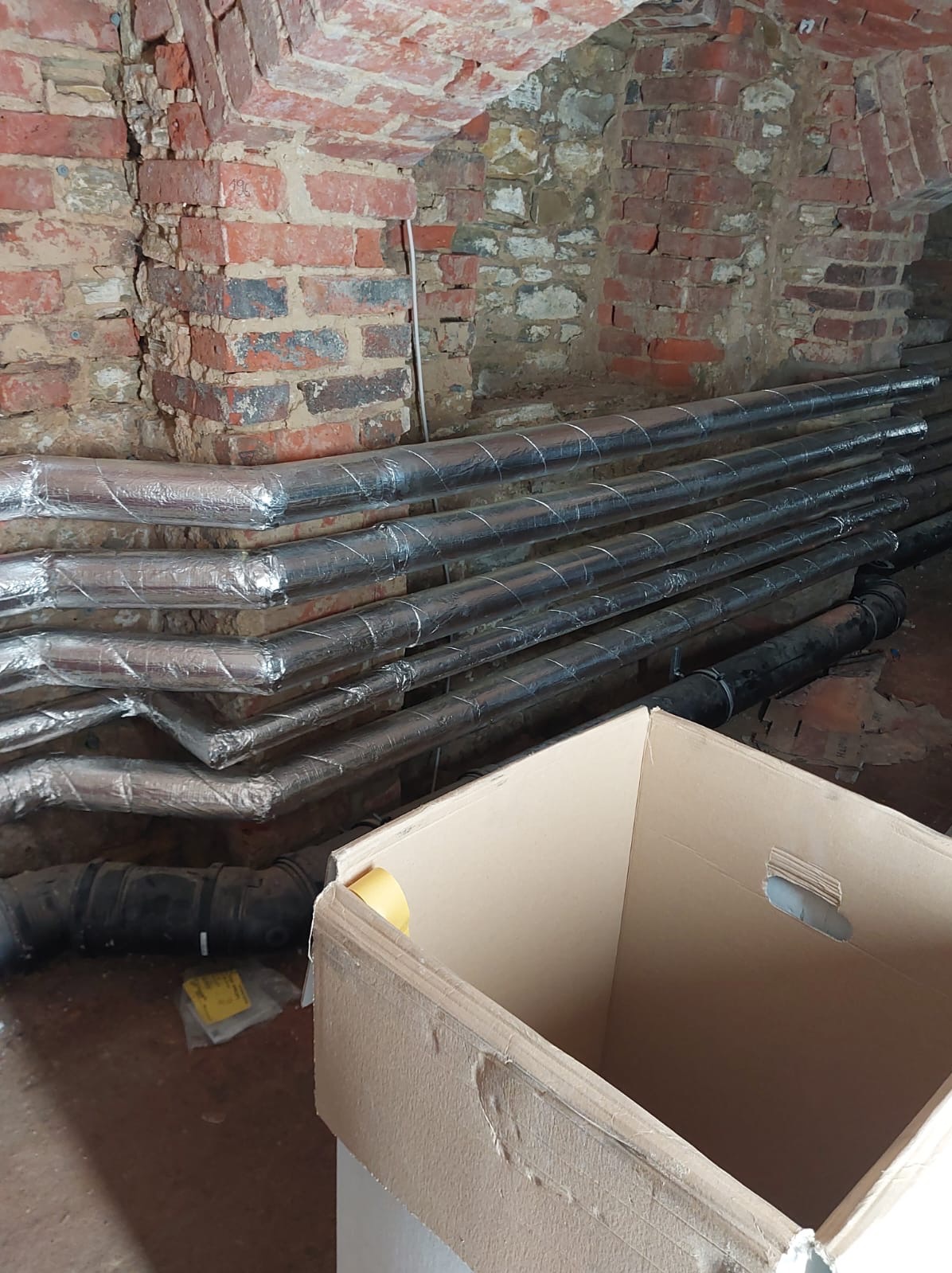 Rury izolowane folią aluminiową ułożone w piwnicy z ceglanym sklepieniem, obok kartonowe pudło. Widoczne złącza rur. Prace instalacyjne w trakcie realizacji.