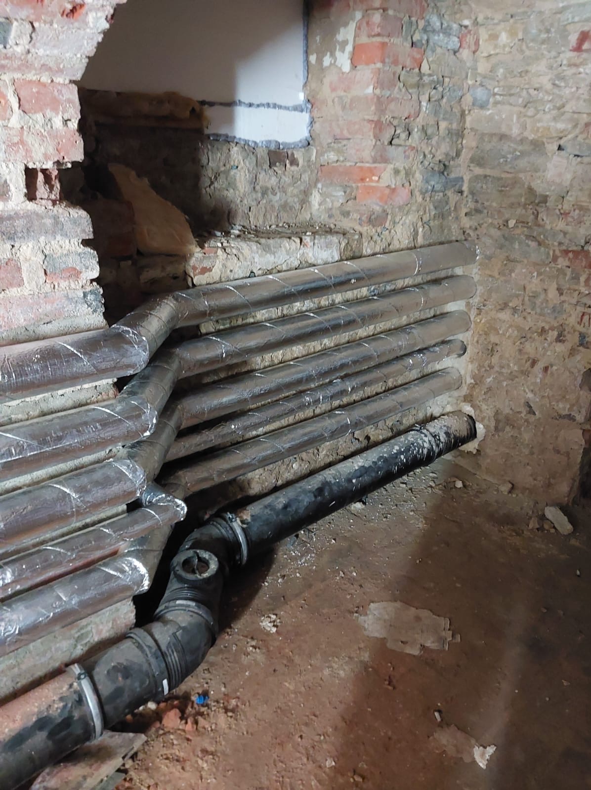 Rury instalacji grzewczej, zaizolowane folią aluminiową, ułożone w surowym, ceglanym wnętrzu. Widoczne czarne rury kanalizacyjne i fragment ściany z ubytkiem tynku.