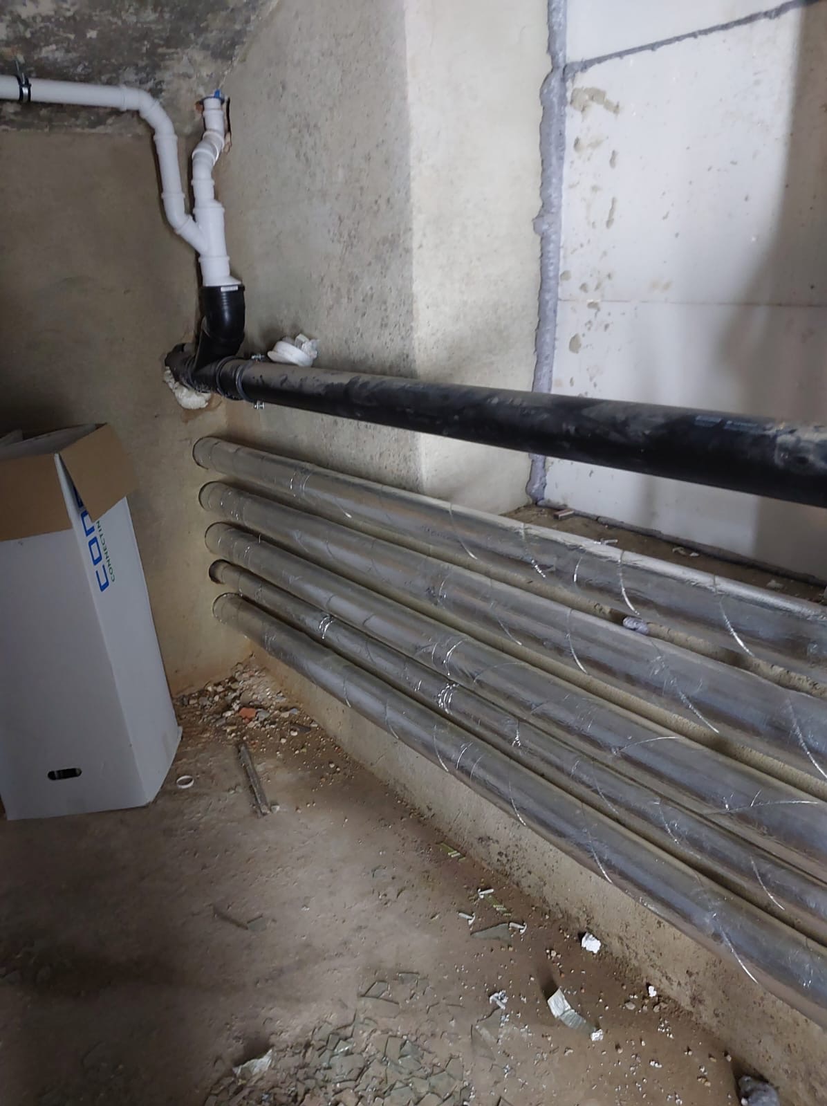 Rury instalacyjne, czarne i srebrne, ułożone wzdłuż ściany w surowym wnętrzu. Widoczne łączenia i elementy instalacji, kartonowe pudło w tle. Surowy stan wykończenia.