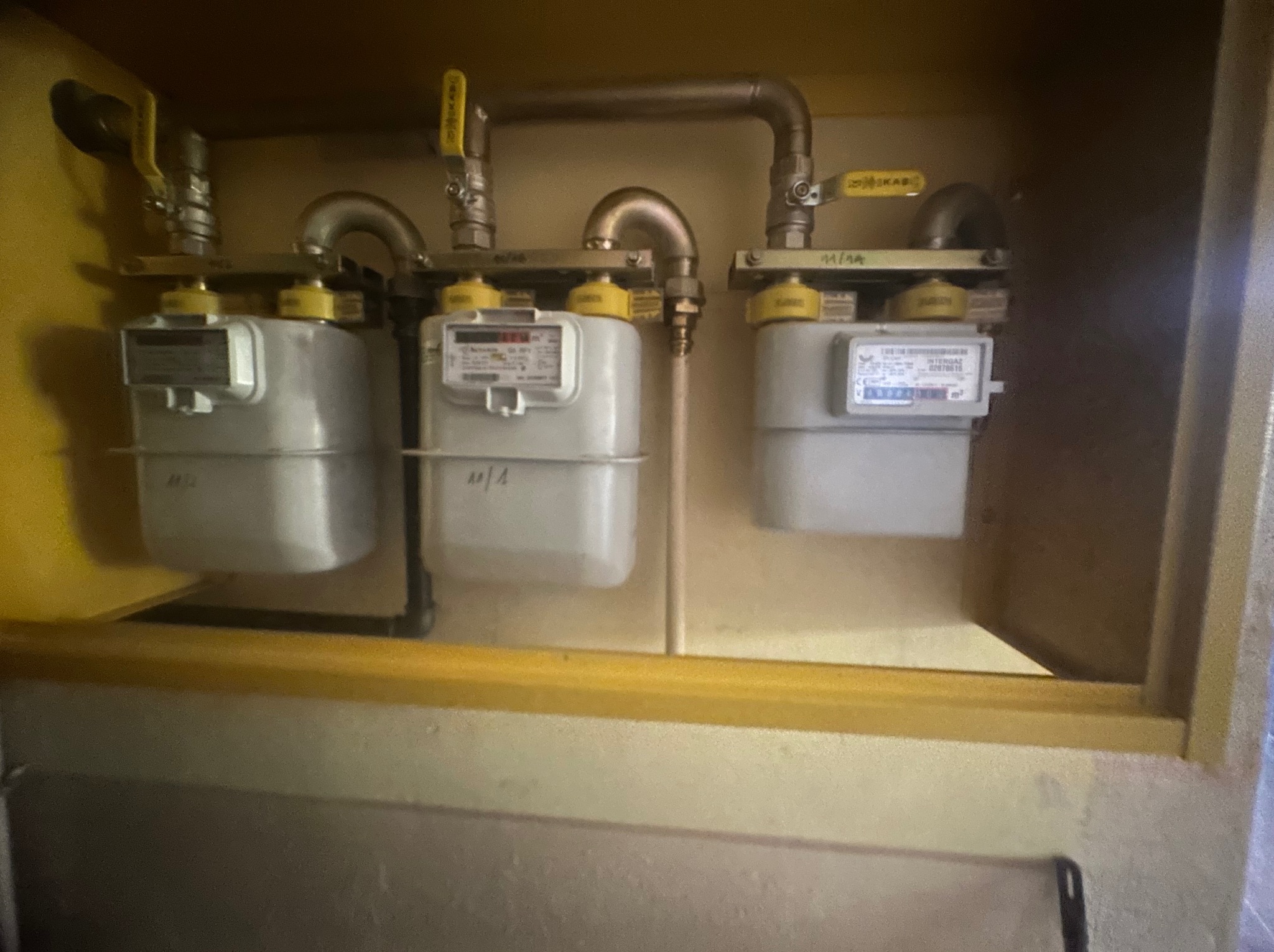 Trzy gazomierze w żółtej szafce, połączone rurami z żółtymi zaworami, widoczne oznaczenia '11/1A' i '11/1', oświetlenie z góry.