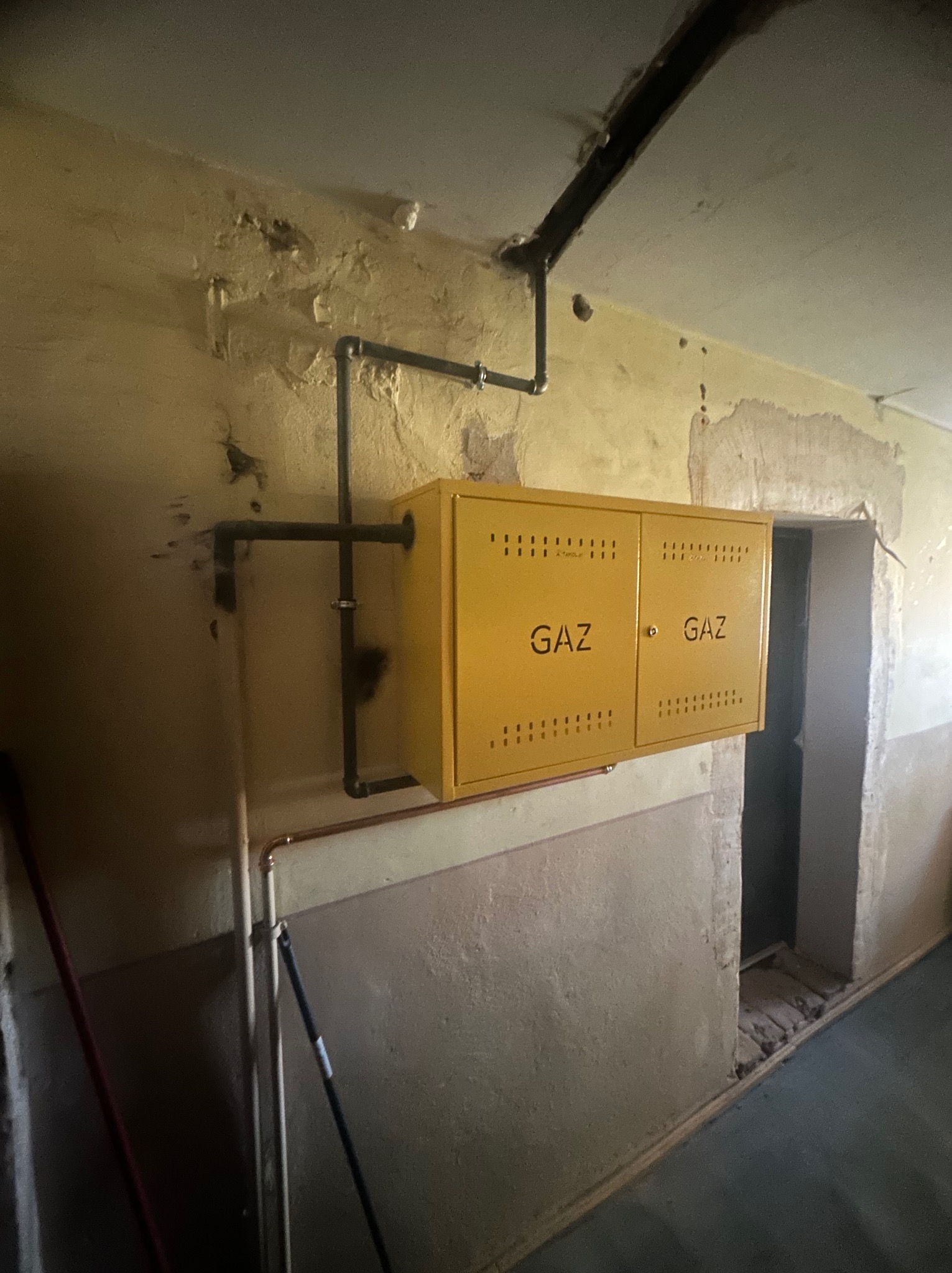 Żółta szafka gazowa z napisem 'GAZ' na ścianie z widocznymi rurami instalacji gazowej. Widoczne ślady remontu.