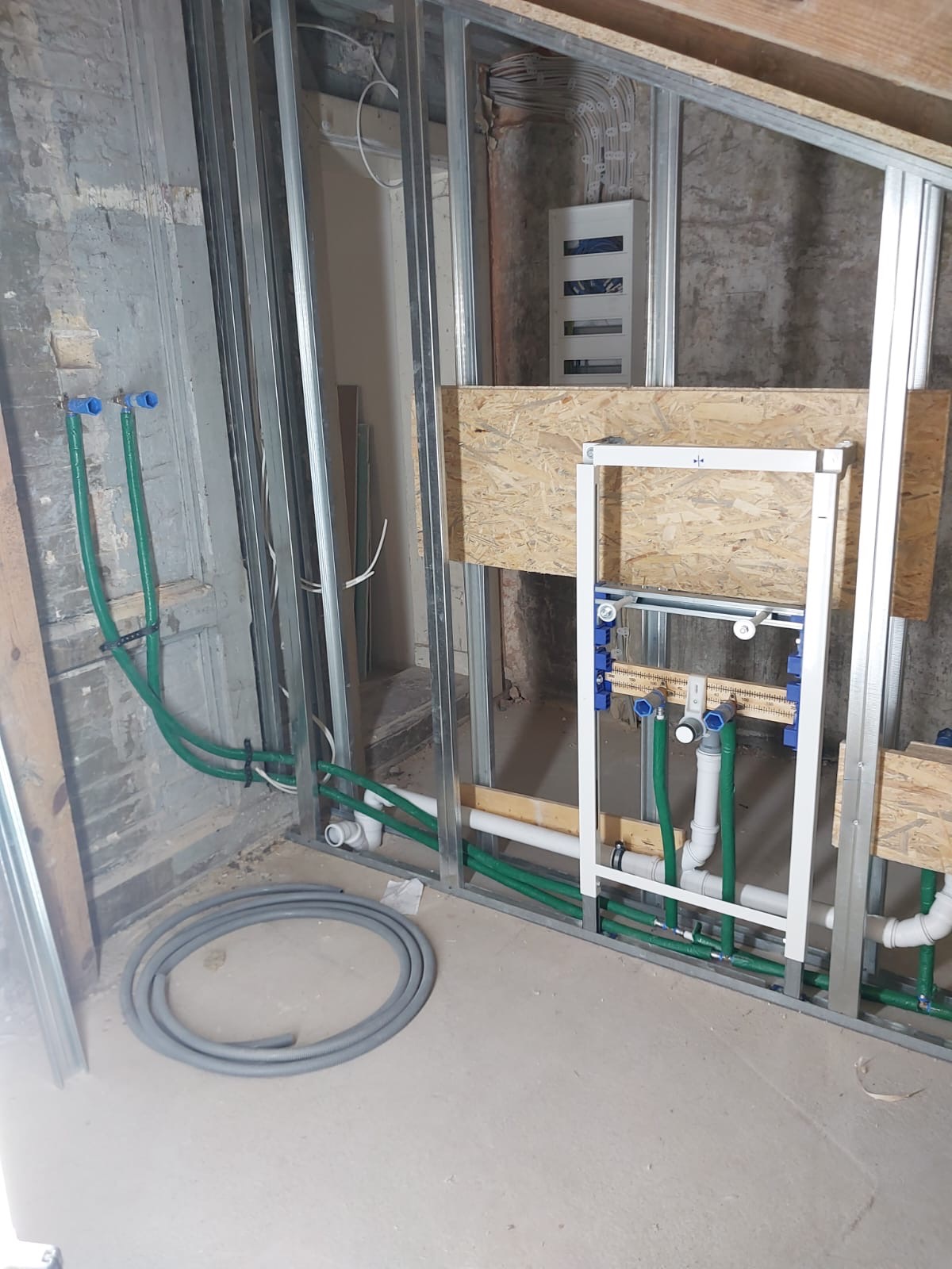 Wnętrze w trakcie remontu: metalowa konstrukcja, instalacja hydrauliczna z zielonych rur, białe rury kanalizacyjne i szara rura karbowana na podłodze.