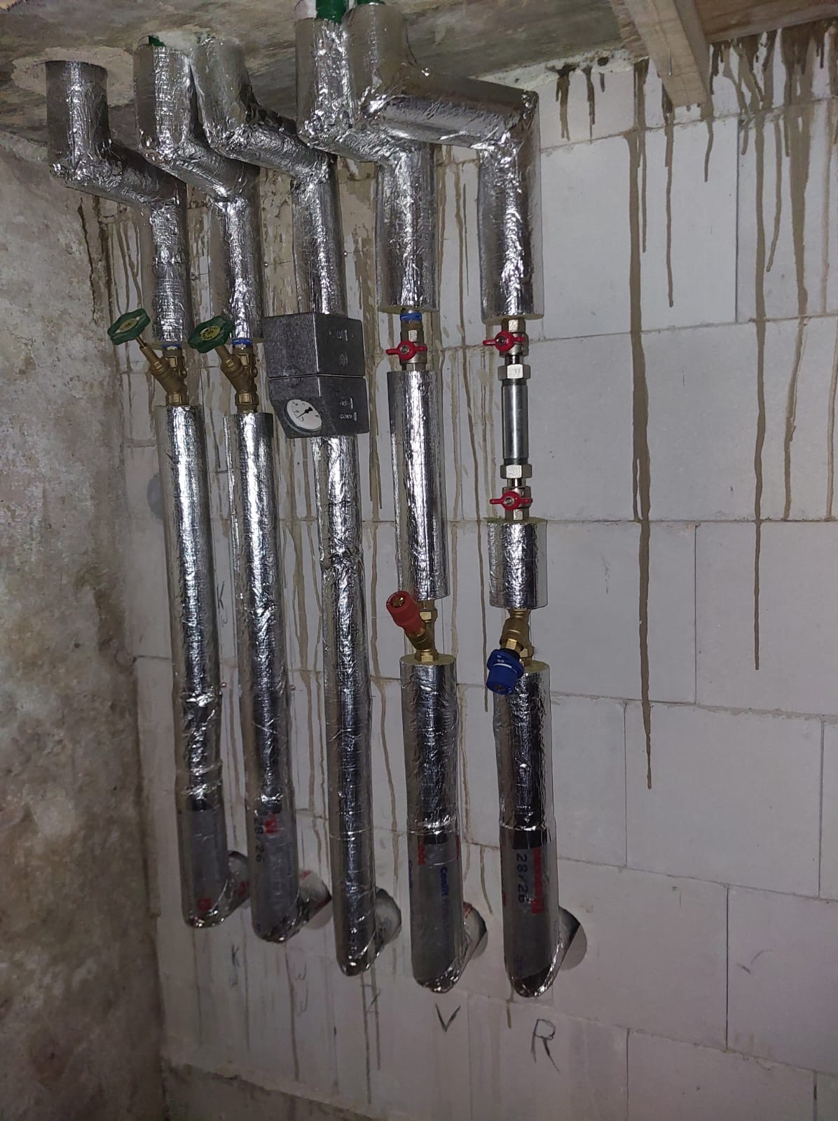 Izolowane rury grzewcze z zaworami i manometrem, zamontowane na ścianie z bloków betonowych z zaciekami. Detal instalacji grzewczej.