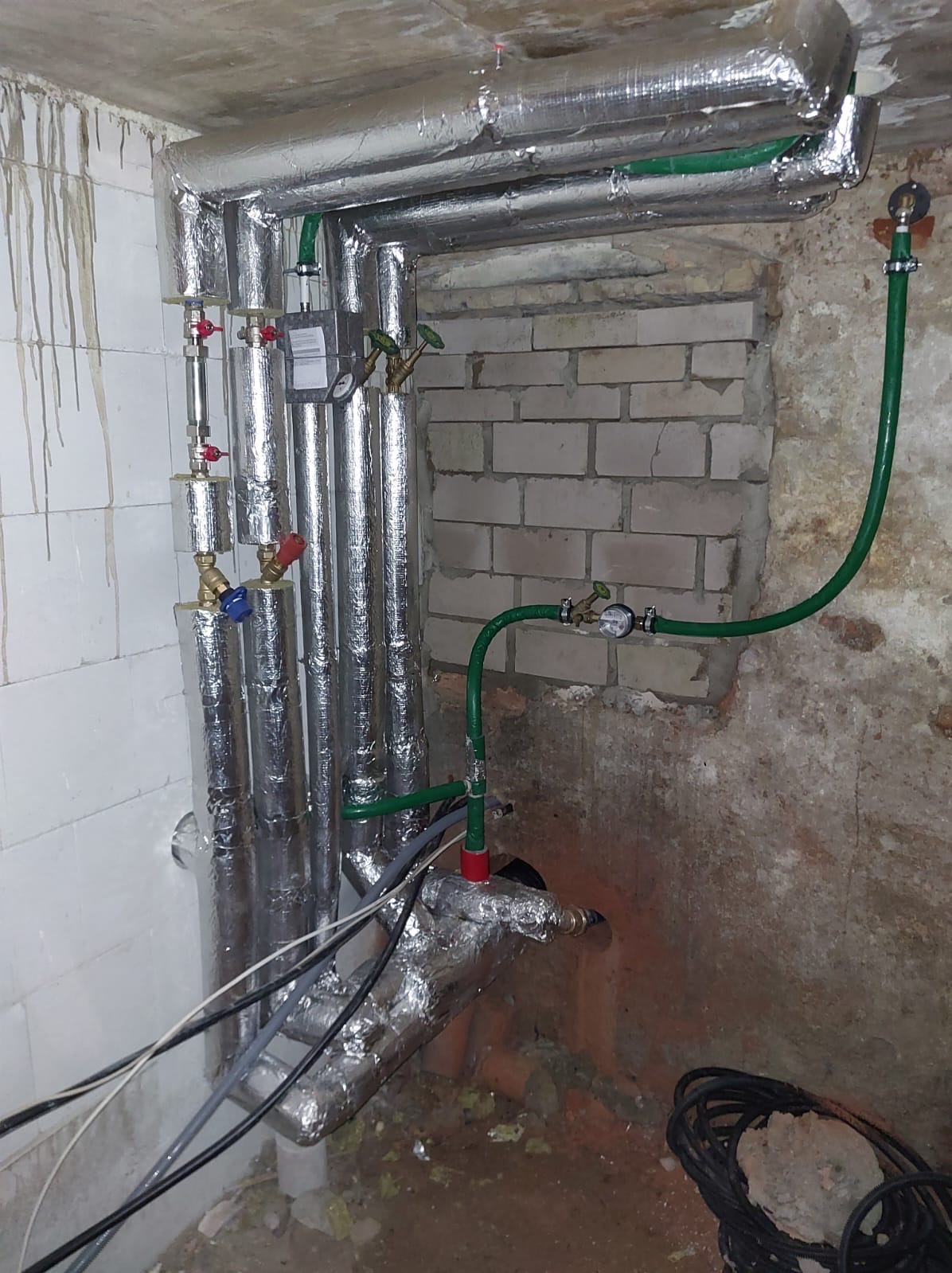 Instalacja grzewcza z rurami w srebrnej izolacji i zielonymi rurami miedzianymi, zaworami i manometrami, zamontowana w piwnicy z surowymi ścianami.