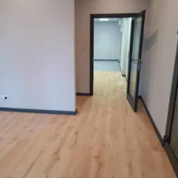 Jasne panele podłogowe w nowym biurze, widok na otwarte drzwi z czarną futryną i szare ściany z listwami przypodłogowymi w kolorze antracytu, fragment gniazdka elektrycznego po lewej stronie.