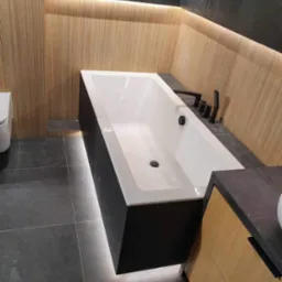 Nowoczesna łazienka z podświetlaną wanną obudowaną czarnym panelem, drewnianą ścianą z ukrytym oświetleniem LED i ciemnymi płytkami podłogowymi.