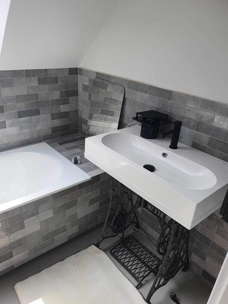 Biała umywalka o owalnym kształcie, zamontowana na odrestaurowanej, żeliwnej podstawie po maszynie do szycia Singer, w łazience wykończonej szarą, cegiełkową płytką, z białą wanną i lustrem opartym...