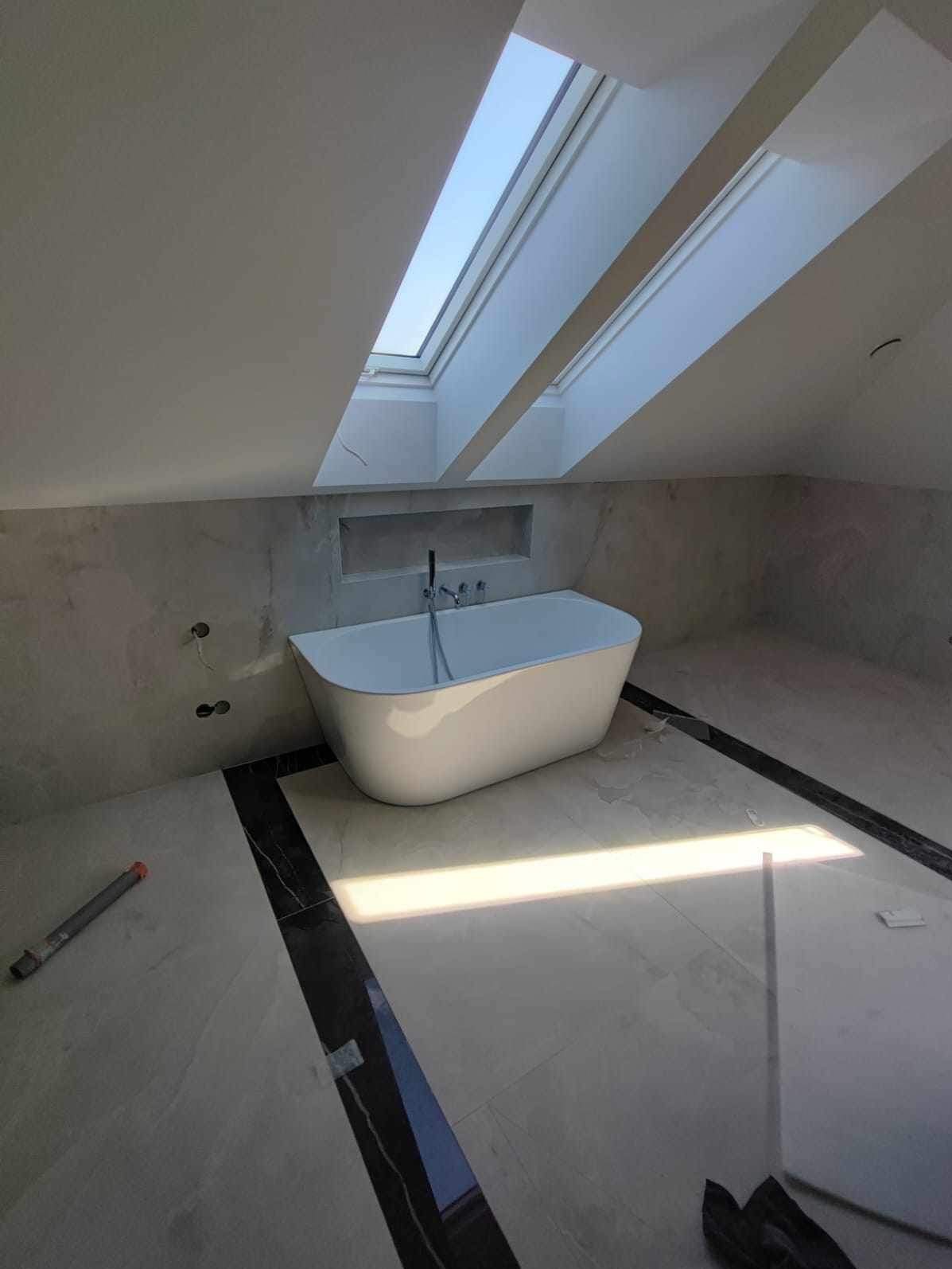 Nowoczesna łazienka na poddaszu z wolnostojącą wanną pod oknem dachowym, szare płytki imitujące marmur na ścianach i podłodze z czarnym akcentem, widoczne niedokończone prace: rurka narzędziowa...