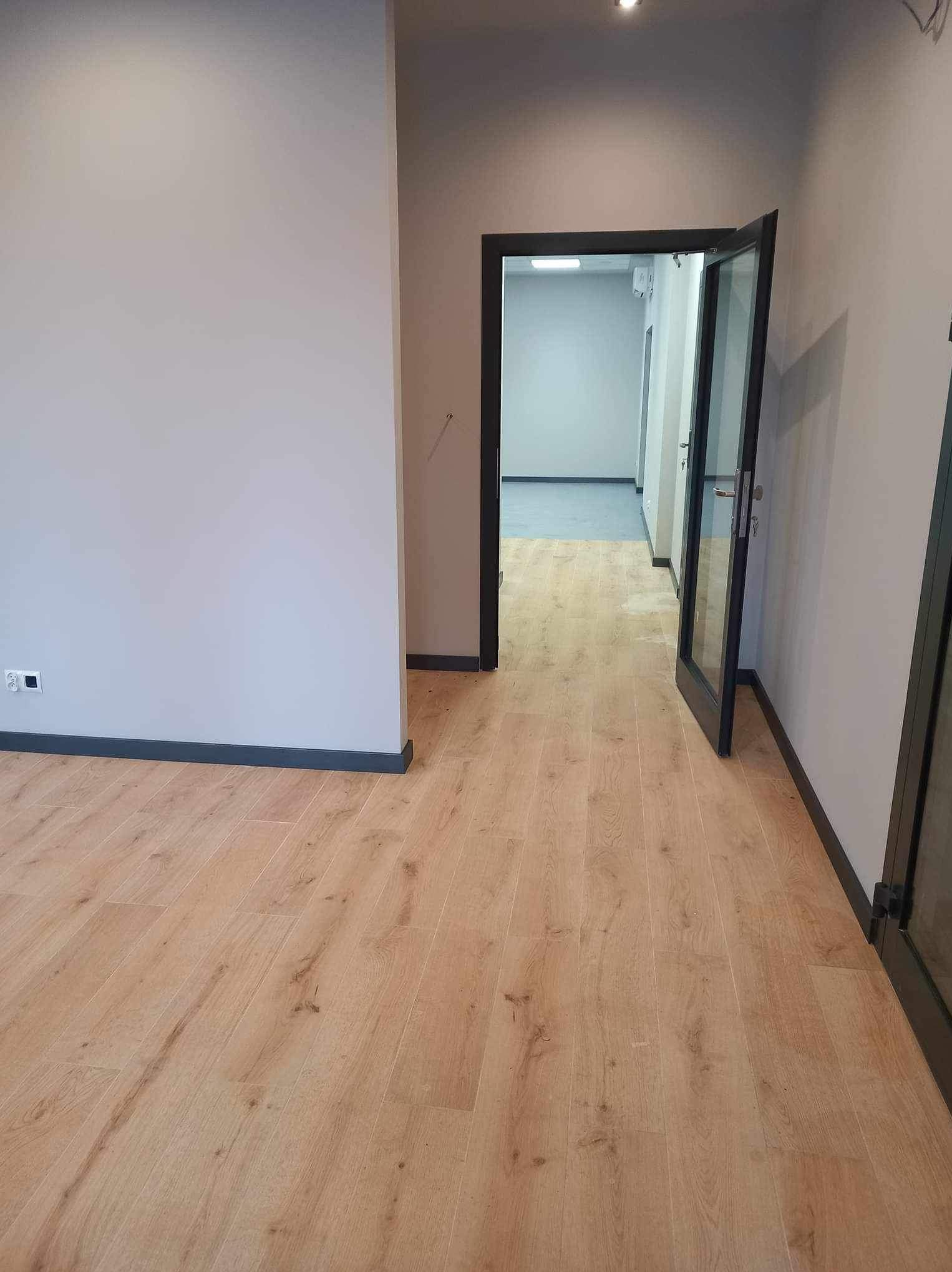 Jasne panele podłogowe w nowym biurze, widok na otwarte drzwi z czarną futryną i szare ściany z listwami przypodłogowymi w kolorze antracytu, fragment gniazdka elektrycznego po lewej stronie.