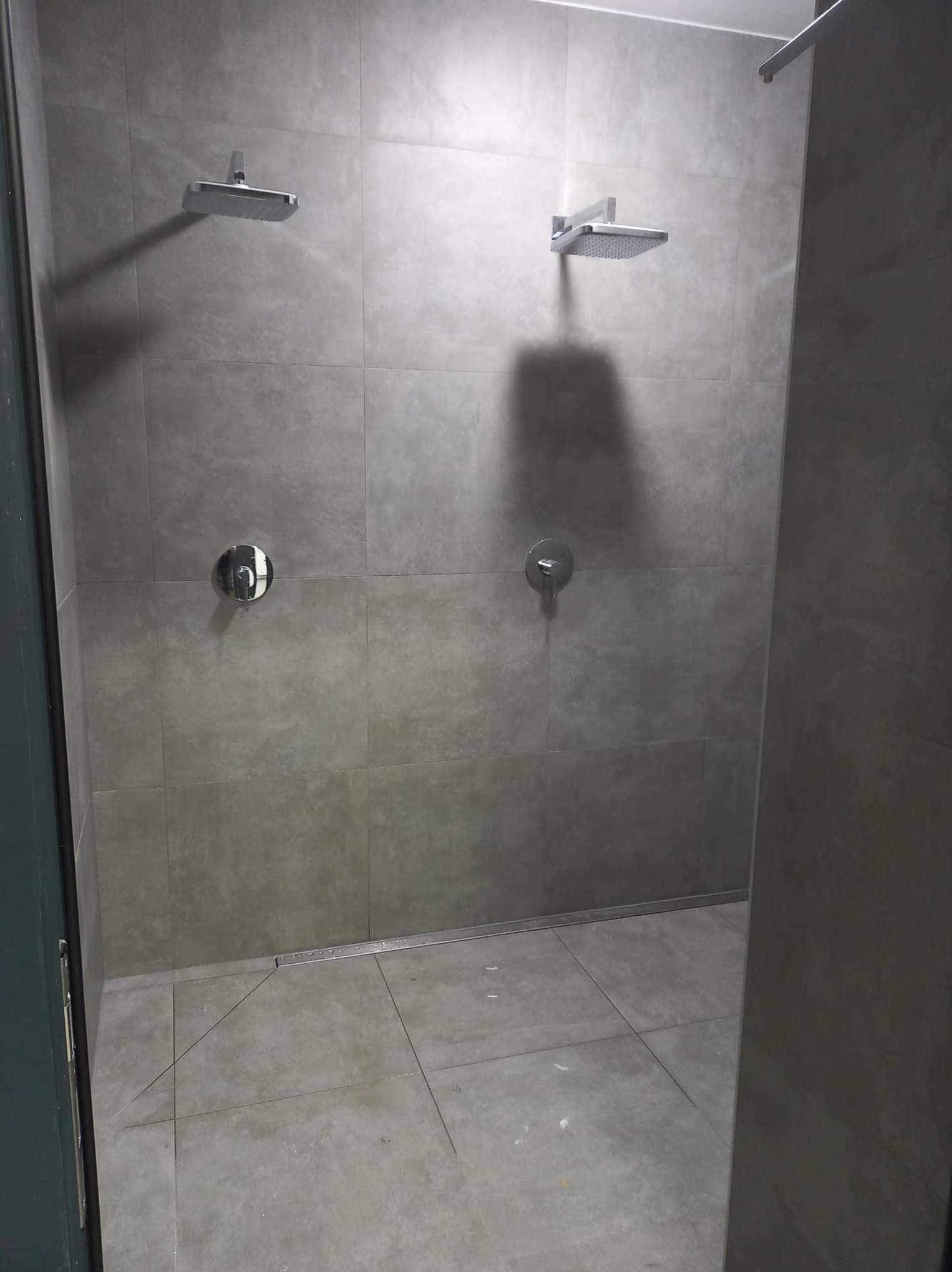 Wykończona łazienka z dwoma prysznicami typu deszczownica, szare płytki na ścianach i podłodze, odpływ liniowy w podłodze, widok z boku.