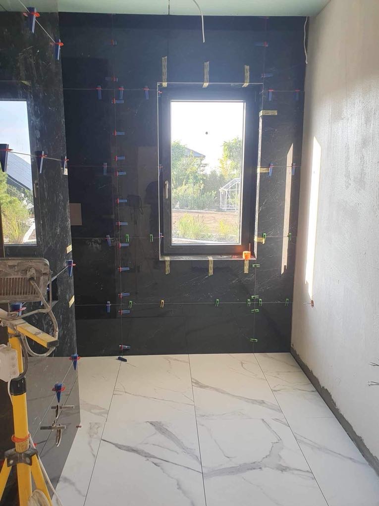 Wnętrze łazienki w trakcie montażu czarnych płytek ściennych i białych płytek podłogowych imitujących marmur, widoczne narzędzia glazurnicze i poziomice krzyżowe oraz okno z widokiem na ogród.
