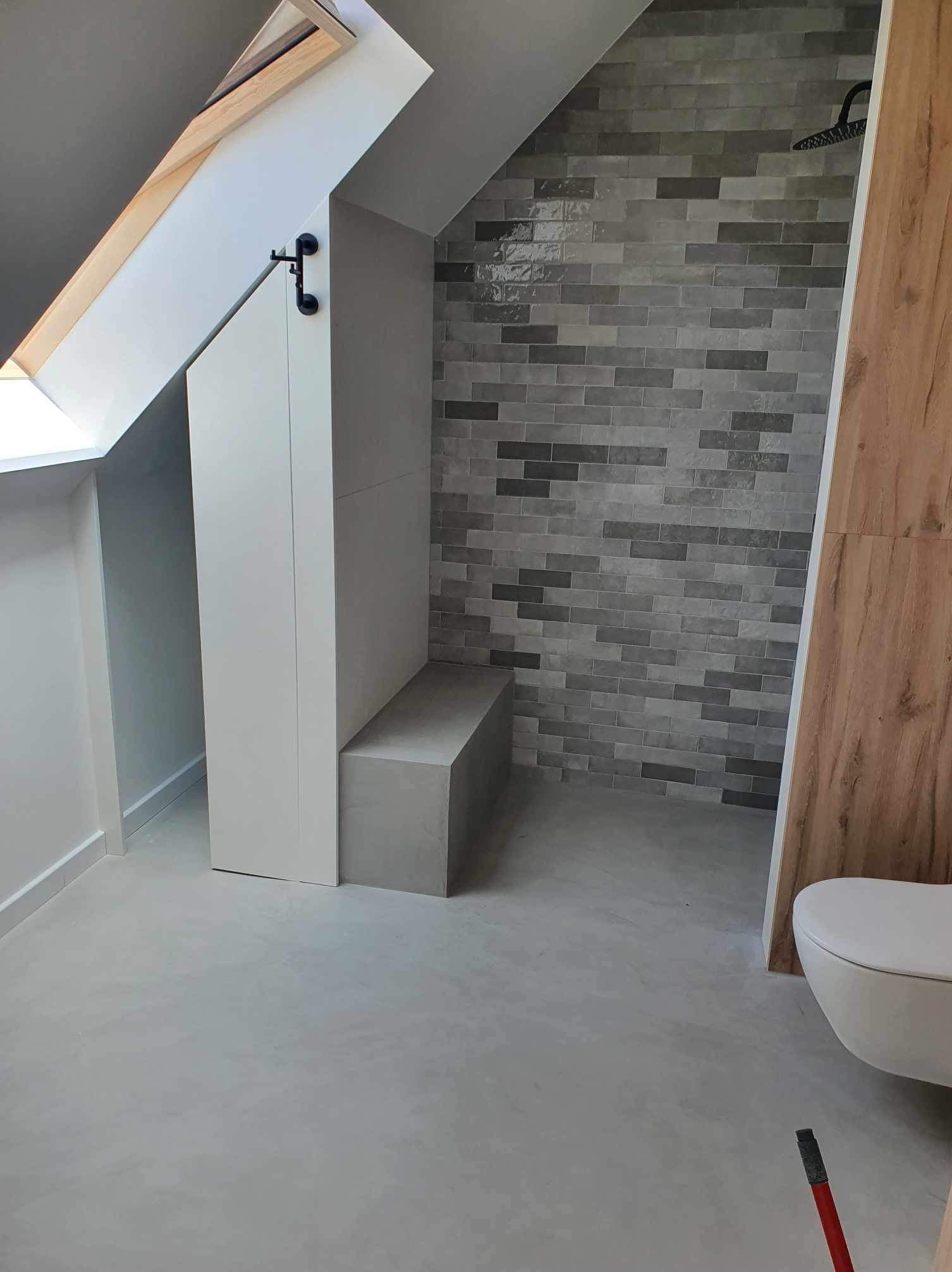 Wykończona łazienka na poddaszu ze skosem, z szarą, ceglastą mozaiką pod prysznicem, zabudowaną szafką z siedziskiem i podwieszaną toaletą. Widoczny fragment okna dachowego.