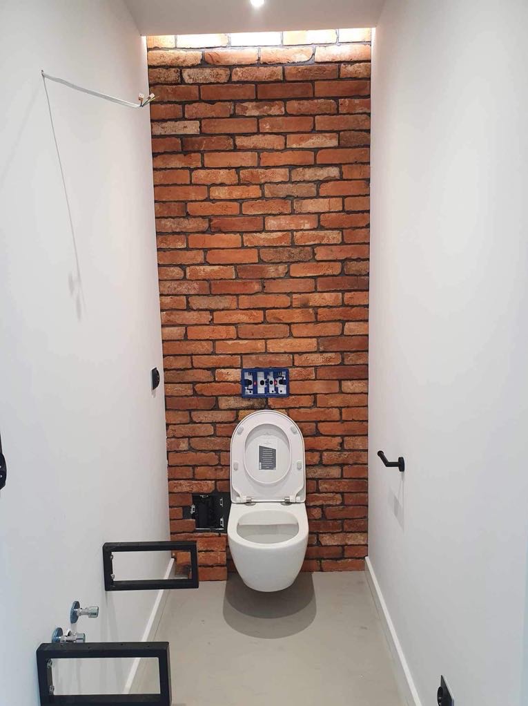 Wąska toaleta we Wrocławiu z białą wiszącą muszlą klozetową na tle ściany wyłożonej czerwoną cegłą, widoczne niedokończone podłączenia hydrauliczne i elektryczne.