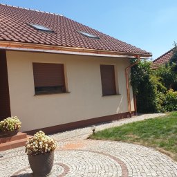 Reymontdach@gmail.com - Dom jednorodzinny z brązową dachówką, miedzianymi rynnami, beżową elewacją i brązowymi roletami zewnętrznymi, widok z poziomu ogrodu z kwiatami w donicach na pierwszym planie.