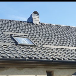 Reymontdach@gmail.com - Nowy dach z szarej blachodachówki z oknem dachowym i kominem wentylacyjnym na tle błękitnego nieba.