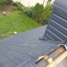 Reymontdach@gmail.com - Montaż szarej blachy trapezowej na dachu z widocznym fragmentem obróbki blacharskiej i osobą w oddali na trawniku.