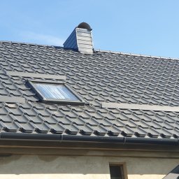 Reymontdach@gmail.com - Grafitowy dach z blachodachówki z oknem dachowym i kominem wentylacyjnym, widok z dołu na tle błękitnego nieba.