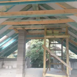 Wnętrze poddasza w trakcie budowy z widoczną konstrukcją drewnianą dachu, oknem dachowym i częściowo wymurowanymi ścianami z betonu komórkowego.