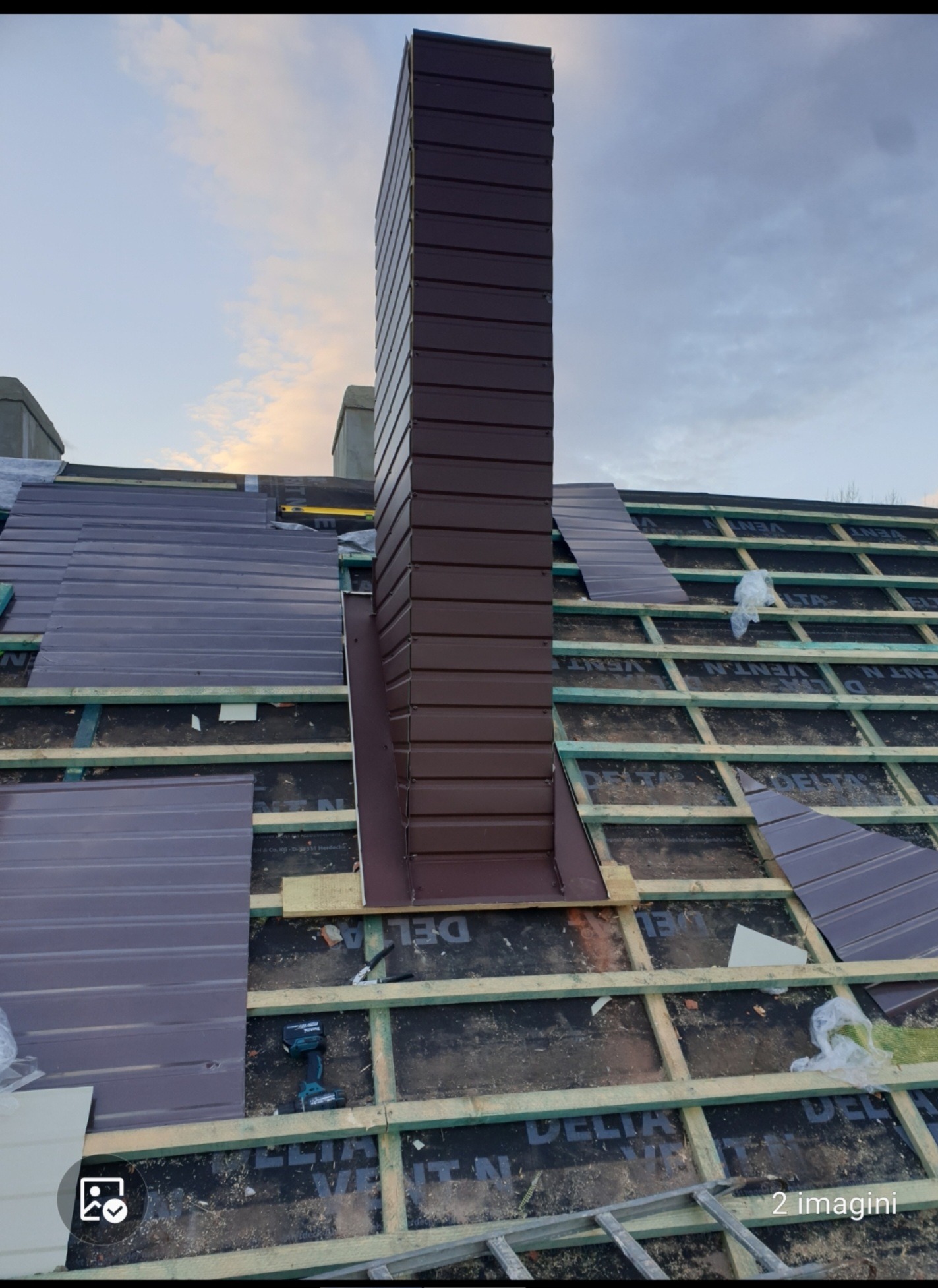 Montaż brązowego komina systemowego na dachu z widoczną konstrukcją drewnianą, częściowo pokrytym blachą w kolorze zbliżonym do komina, w tle pochmurne niebo.