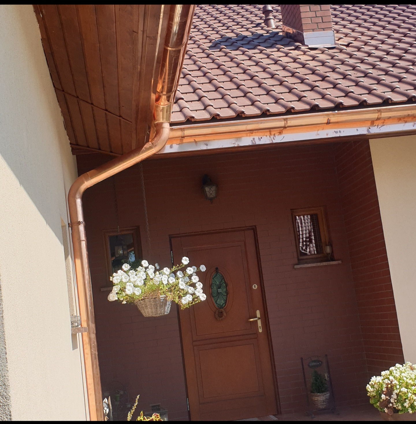 Miedziane orynnowanie domu z brązową dachówką ceramiczną i dekoracjami kwiatowymi przy wejściu.