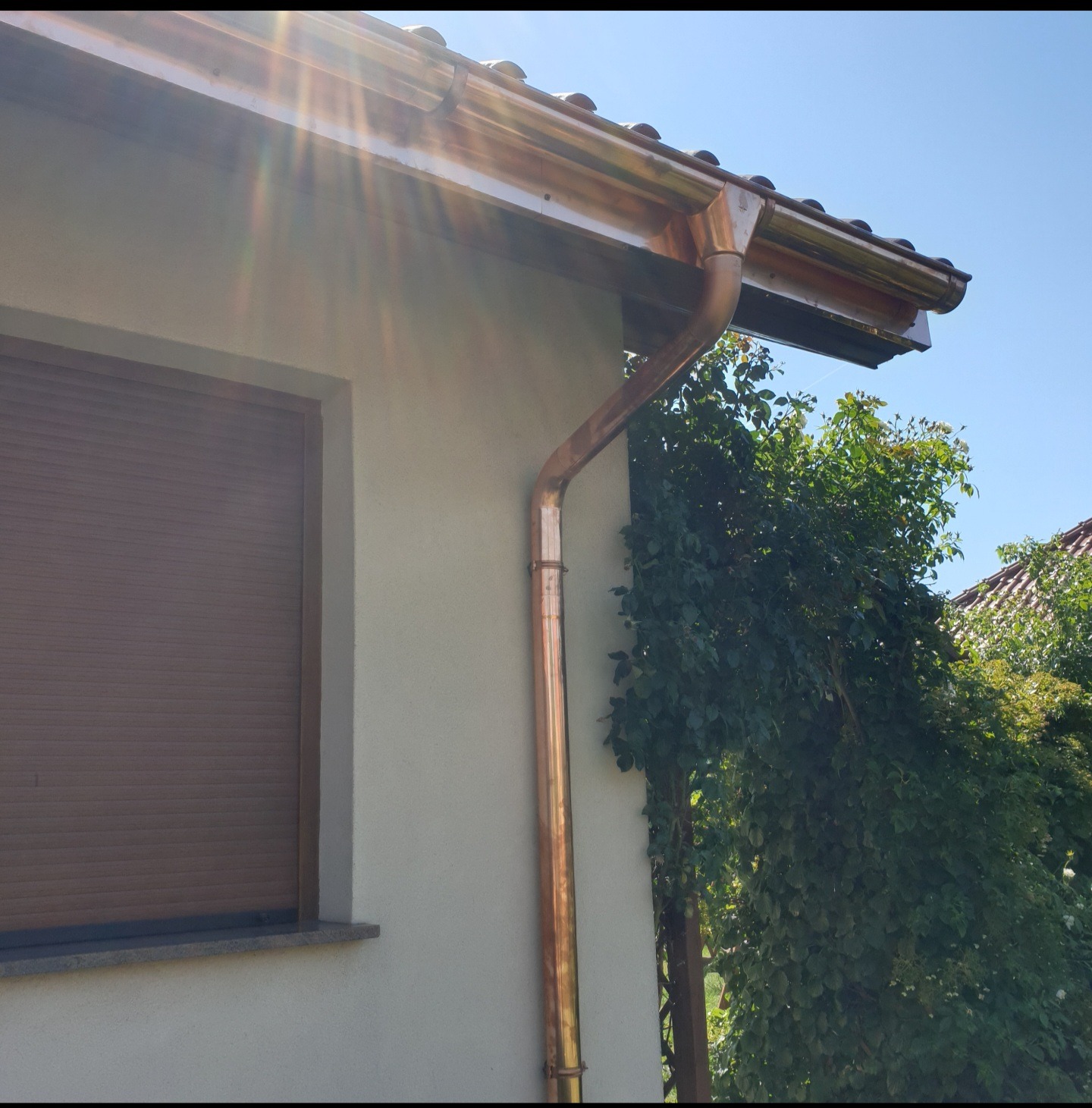 Miedziana rynna spustowa i rynna dachowa zamontowane na elewacji budynku z zamkniętą roletą okienną, widoczne refleksy słońca i zieleń roślin.