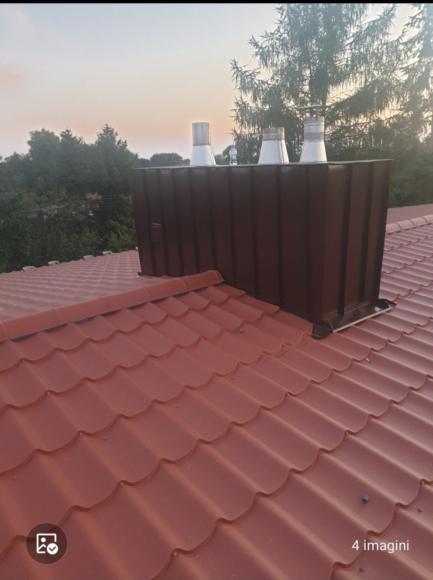 Czerwona dachówka ceramiczna z brązową obróbką komina i trzema metalowymi wkładkami kominowymi na tle drzew i zachodzącego słońca.