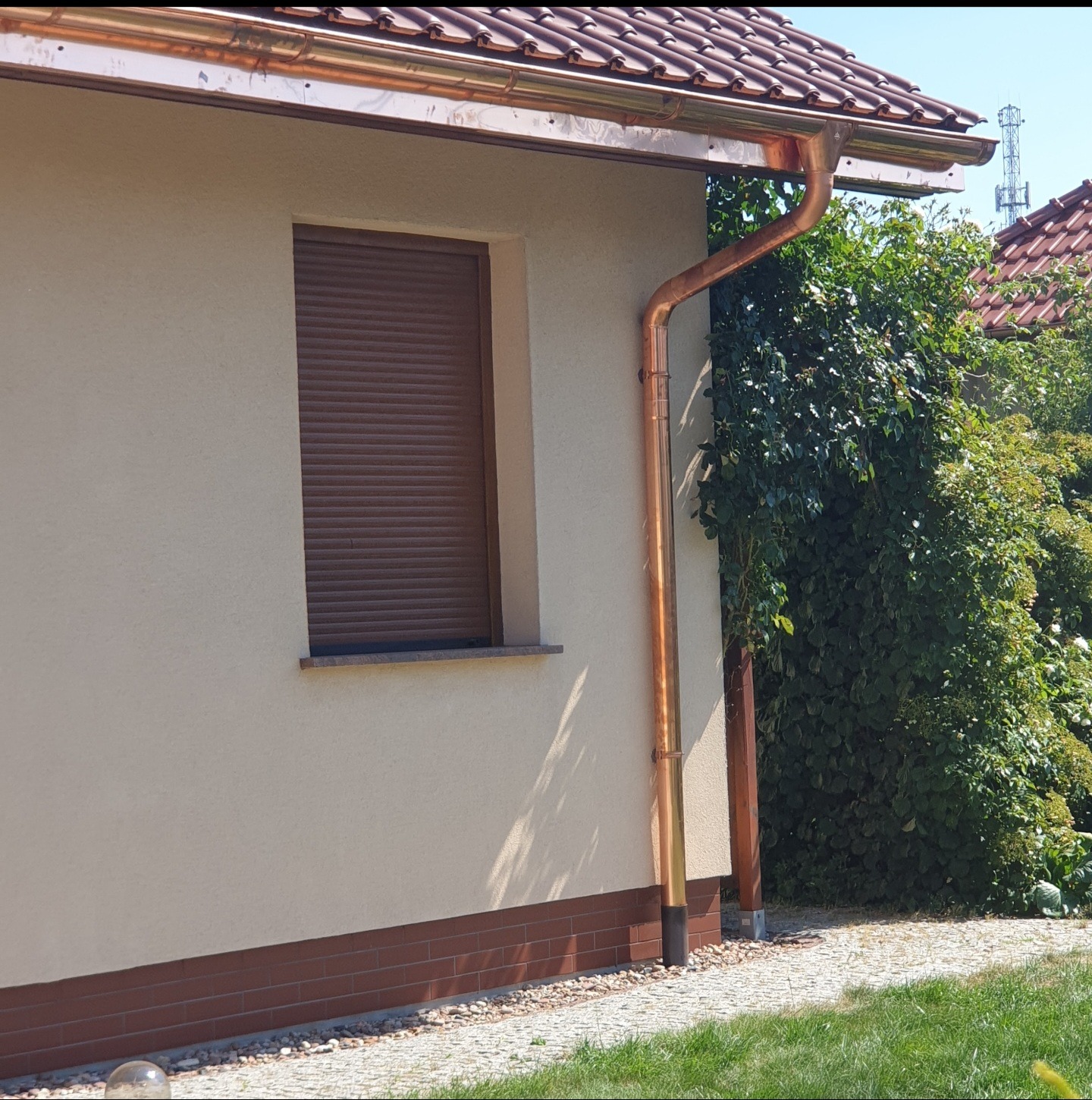 Miedziane rynny pionowe i poziome na elewacji domu z brązową roletą okienną i ceglaną podmurówką, widok z boku.