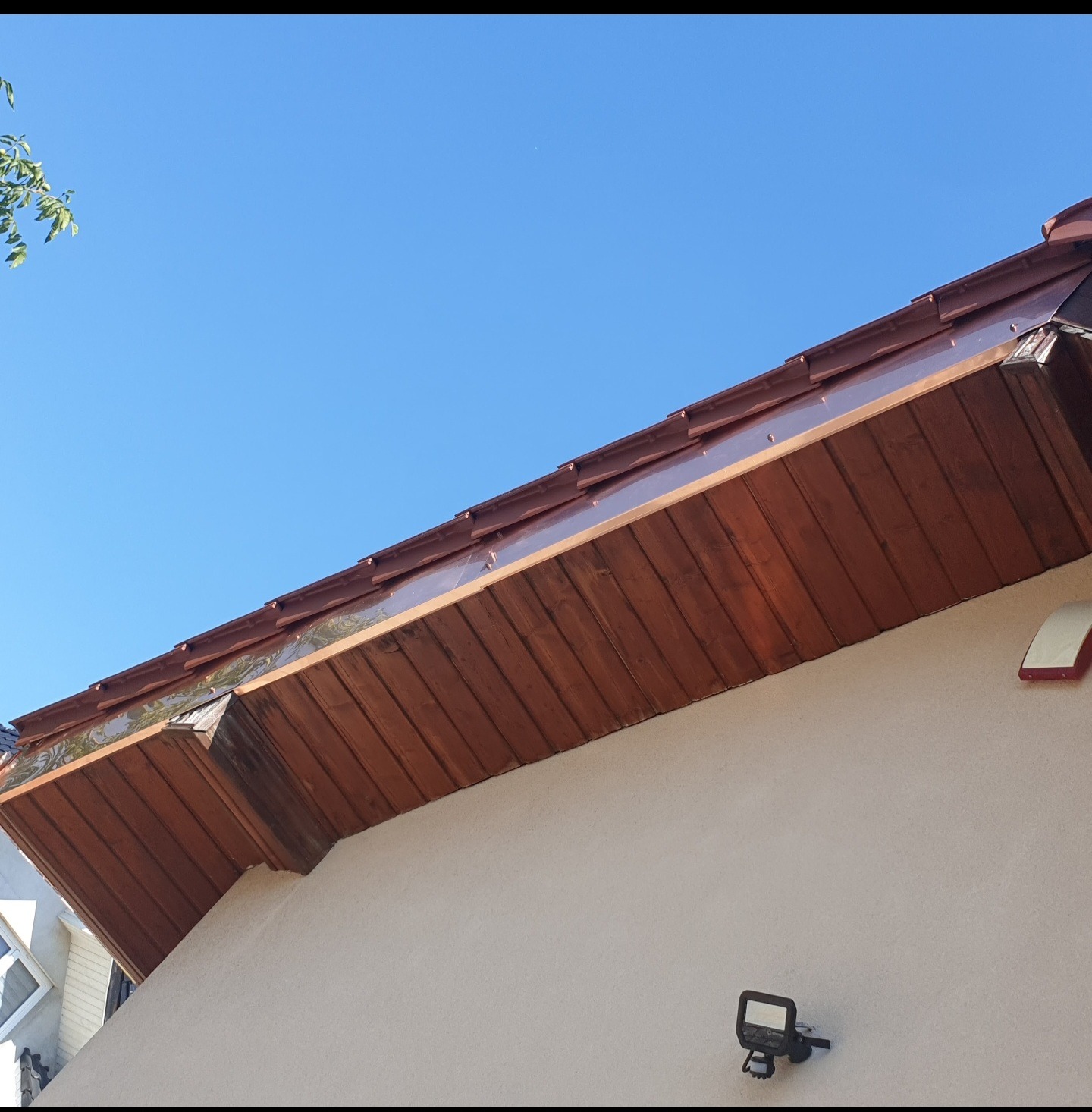 Wykończony okap dachu z brązowymi panelami drewnianymi i błyszczącą metalową obróbką, widok z dołu na tle błękitnego nieba, z fragmentem zieleni drzewa w lewym górnym rogu.