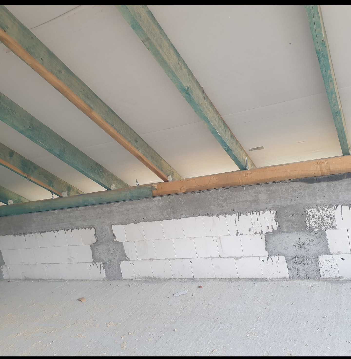 Sufit poddasza w trakcie ocieplania, widoczne drewniane belki, biała izolacja i fragment ściany z bloczków betonowych z widocznymi śladami zaprawy.