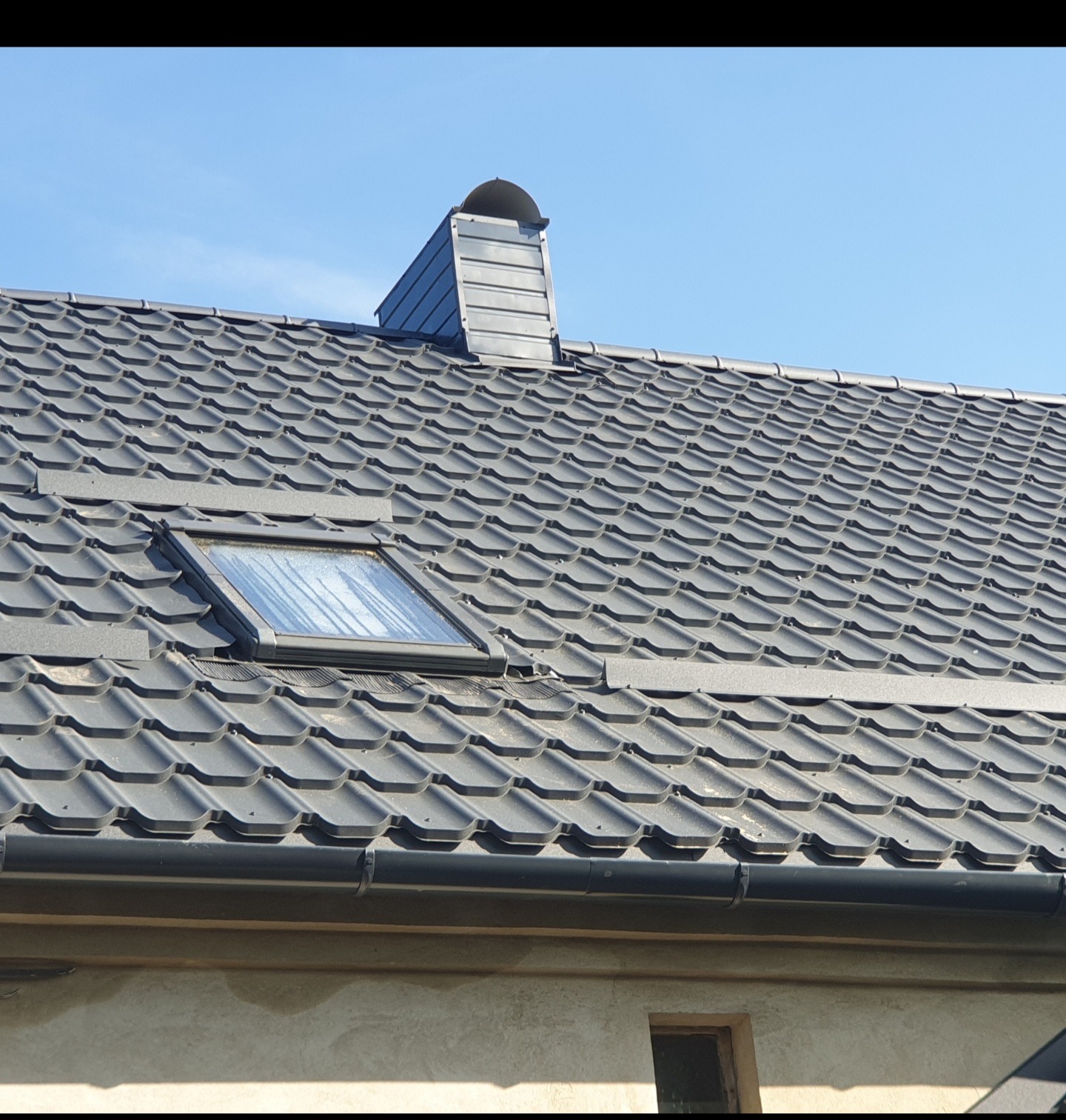 Nowy dach z szarej blachodachówki z oknem dachowym i kominem wentylacyjnym na tle błękitnego nieba.