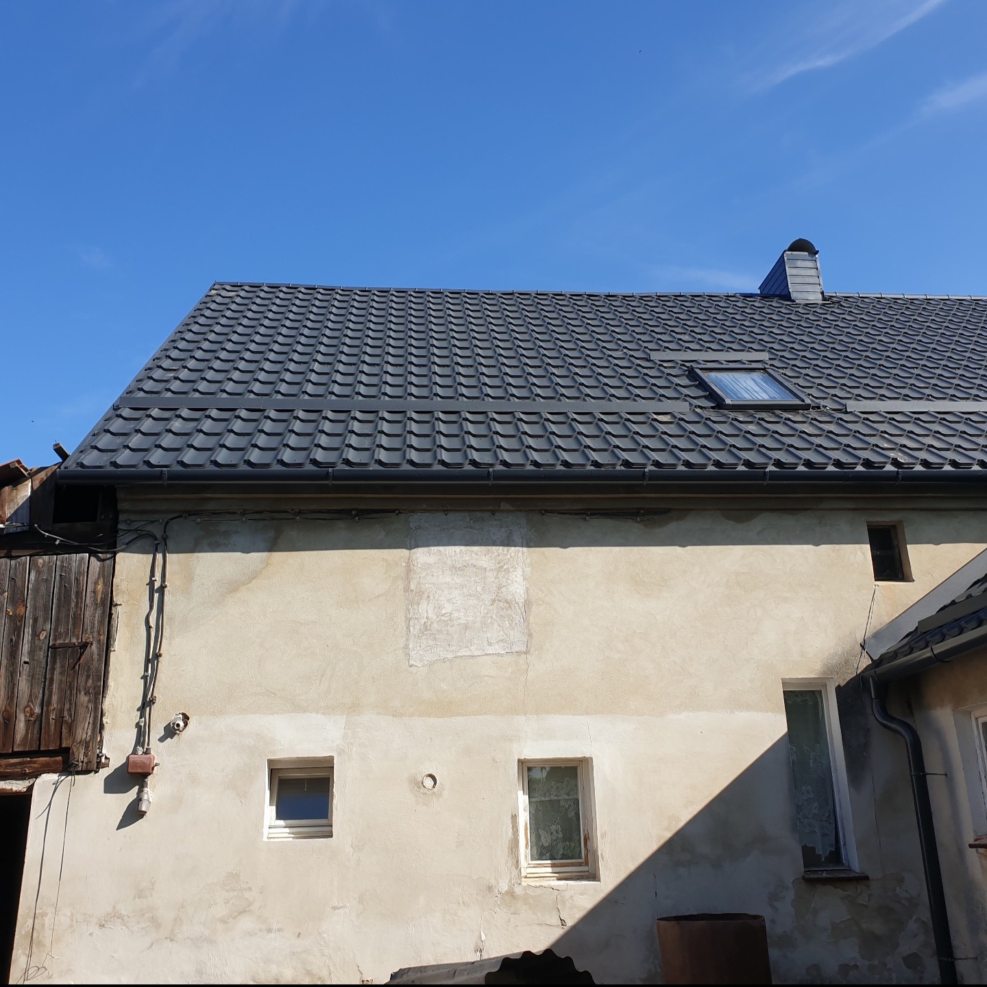 Dom z nowym, ciemnoszarym dachem z dachówki ceramicznej, widoczne okno dachowe i komin, elewacja budynku w odcieniach beżu z małymi oknami.